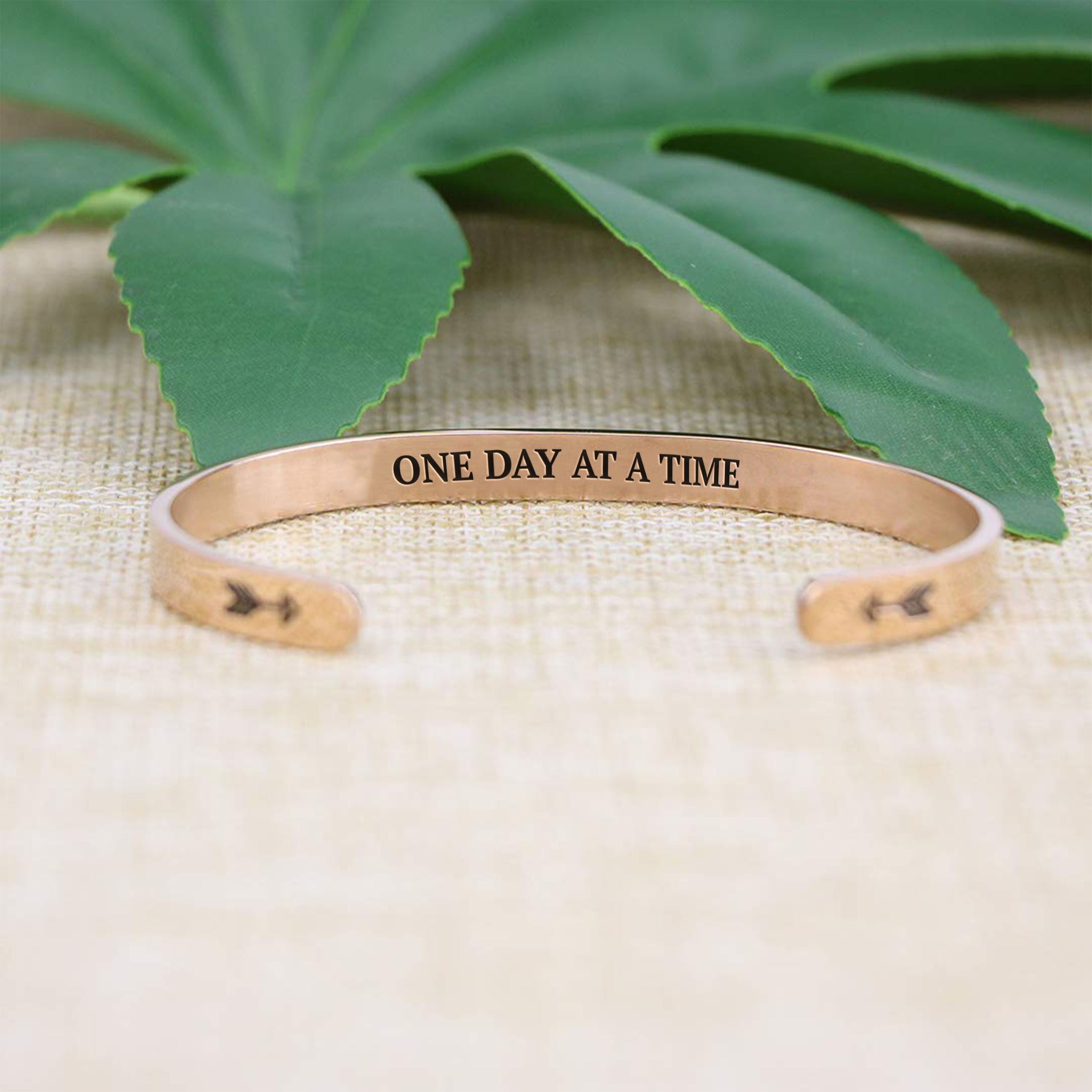 One day at a time bracelet with rose gold plating with message in focus on a burlap surface with a leafy background