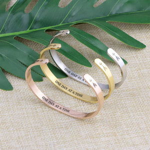 One day at a time bracelets with silver, gold, and rose gold plating standing on a burlap surface with a leafy background