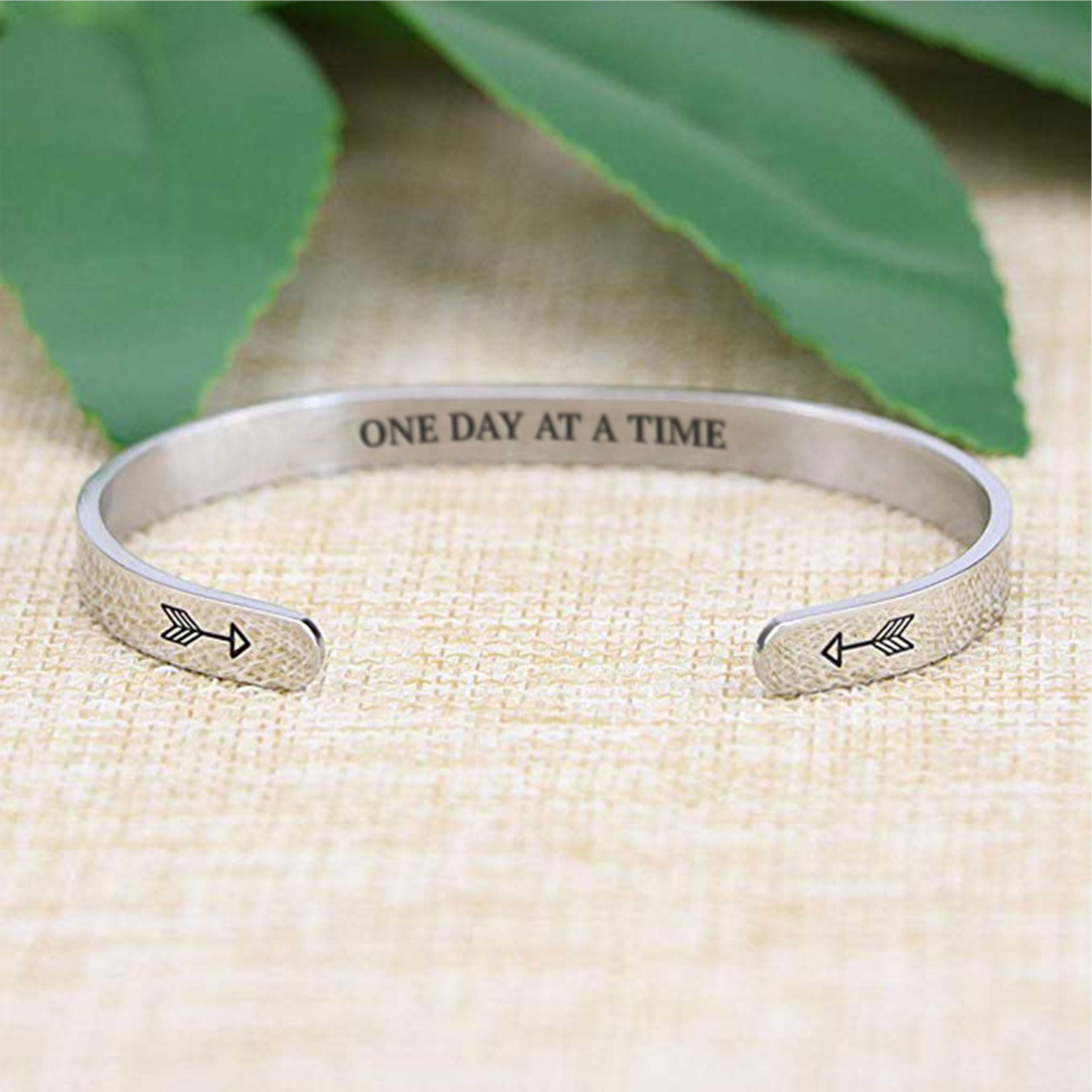 One day at a time bracelet with silver plating with arrows in focus on a burlap surface with a leafy background