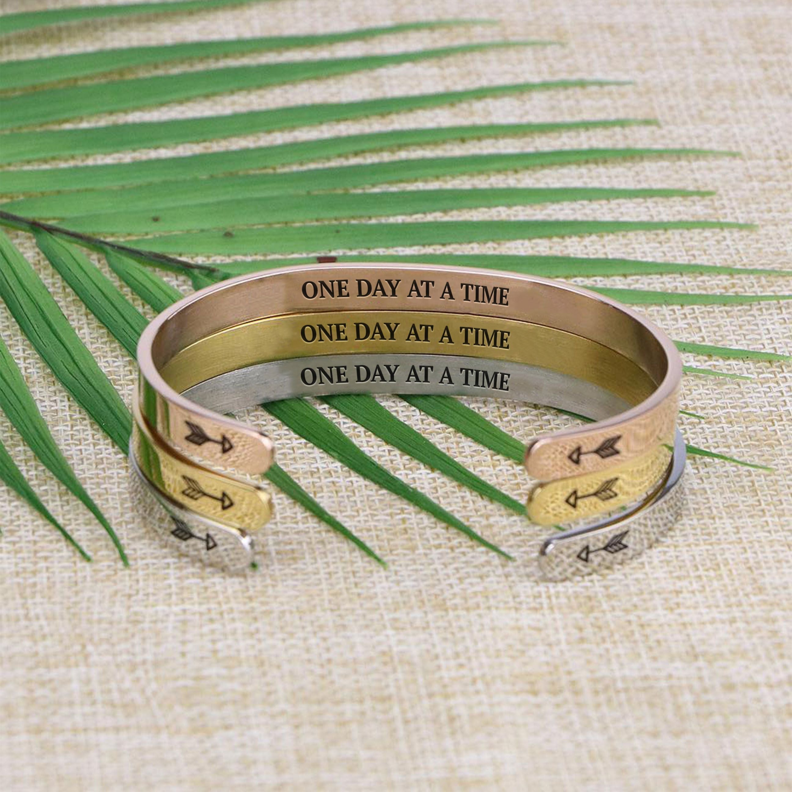 One day at a time bracelets with silver, gold, and rose gold plating stacked on a burlap surface with a leafy background
